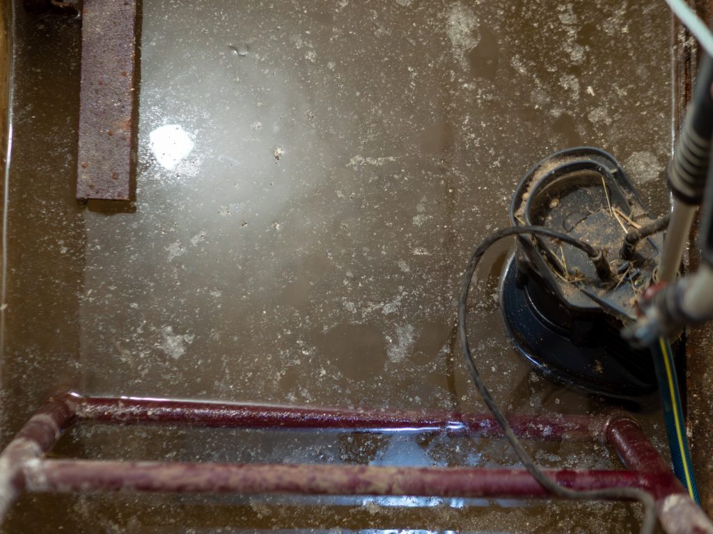 Close up overhead view of black electric submersible water pump working in murky flooded concrete pit with surface bubbles for home basement drainage and flood prevention.