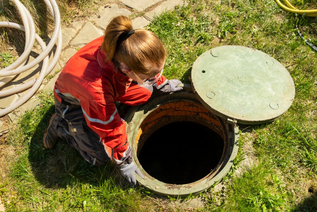 Girl plumber looks into an open draining manhole. Service of the house individual sewerage.